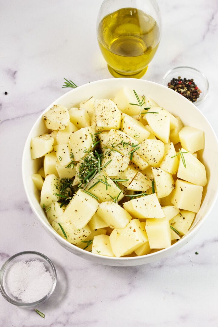 A white bowl filled with cubed roasted rosemary potatoes, seasoned with herbs and pepper, surrounded by a glass of olive oil, a bowl of salt, and a bowl of peppercorns on a white marble surface.