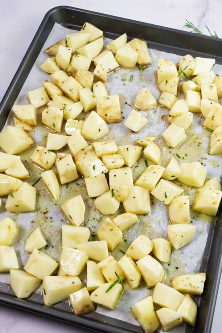 Cubed potatoes tossed with herbs and oil are spread in a single layer on a parchment-lined baking sheet, ready to become perfectly roasted rosemary potatoes.