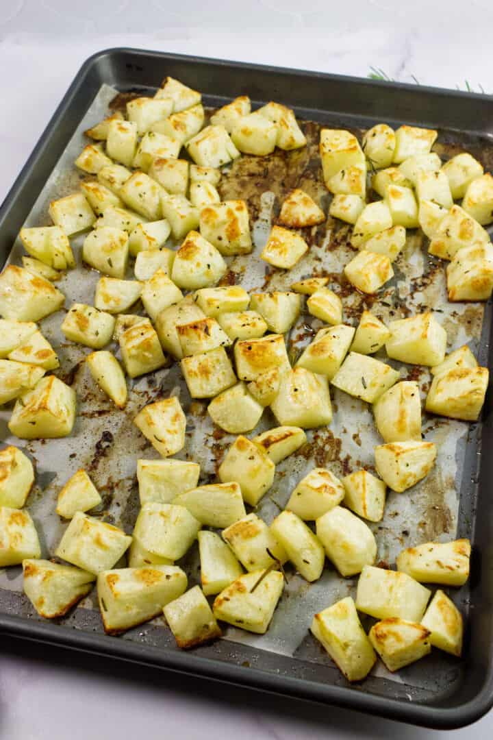 A baking tray filled with evenly spread, golden-brown roasted kohlrabi cubes and roasted rosemary potatoes on parchment paper.