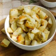 A white bowl filled with roasted rosemary potatoes sits on a wooden surface, with some herbs sprinkled on the golden-brown, seasoned cubes.