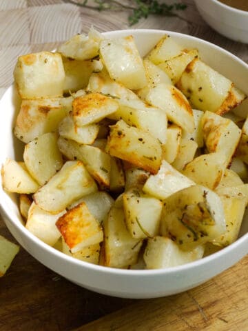 A white bowl filled with roasted rosemary potatoes sits on a wooden surface, with some herbs sprinkled on the golden-brown, seasoned cubes.