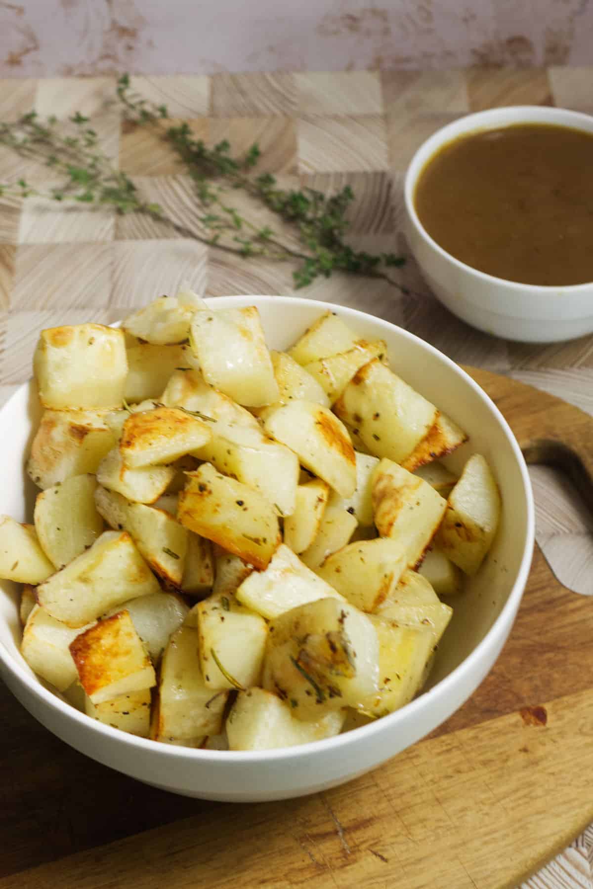 A white bowl filled with roasted rosemary potatoes sits on a wooden cutting board, with a sprig of herbs and a small bowl of brown gravy in the background.