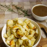 A white bowl filled with golden roasted rosemary potatoes sits on a wooden board. In the background, a small white bowl of brown gravy and sprigs of fresh herbs are visible.