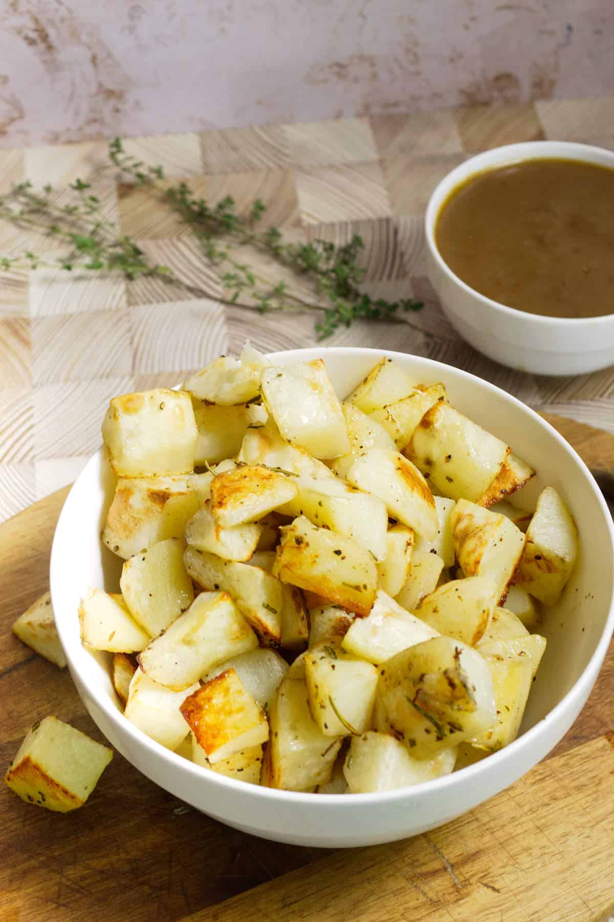 A white bowl filled with roasted rosemary potatoes sits on a wooden board. Fresh herbs are scattered in the background, and a small bowl of brown gravy is placed to the side.