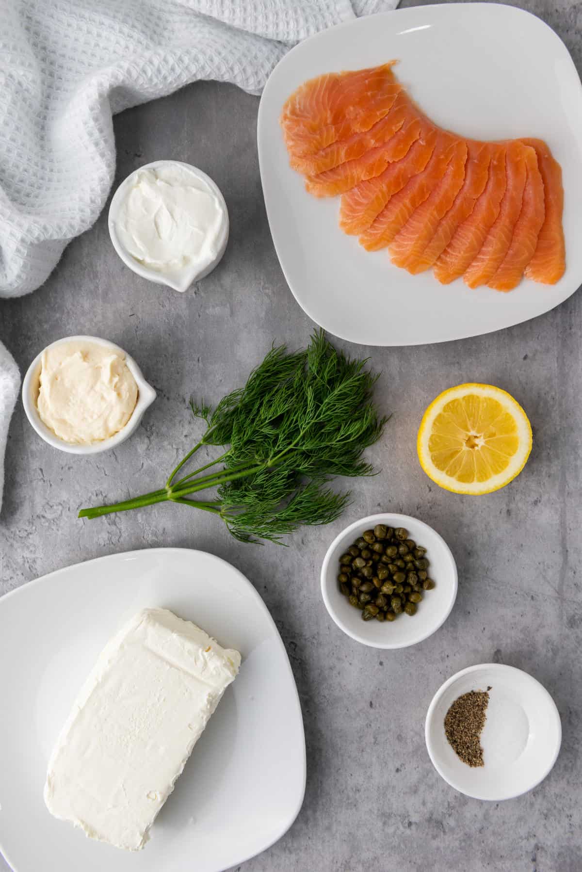 Overhead view of smoked salmon dip ingredients—lox, cream cheese, fresh dill, half a lemon, capers, black pepper, and a sauce in small bowls on a gray surface, with a white towel partially visible in the corner.