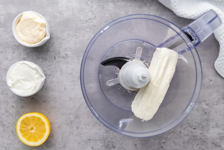 A food processor bowl with a block of cream cheese, next to small bowls of mayonnaise and sour cream, half a lemon, all on a gray countertop-perfect ingredients for making smoked salmon dip.