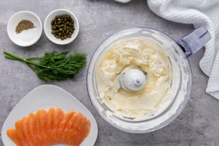A food processor with creamy smoked salmon dip sits on a countertop, surrounded by fresh dill, smoked salmon slices, a small bowl of capers, and pepper and salt, with a white textured towel nearby.