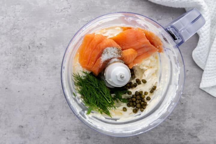 A food processor bowl contains cream cheese, smoked salmon slices, fresh dill, capers, and a sprinkle of salt, ready to be blended into a delicious smoked salmon dip. The processor sits on a gray countertop beside a white textured towel.