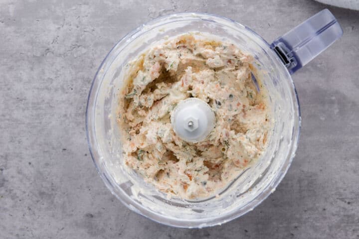A food processor bowl filled with creamy smoked salmon dip, mixed with visible herbs and small bits of ingredients, sits on a gray countertop.