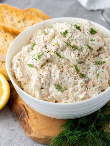 A white bowl filled with creamy smoked salmon dip, garnished with fresh dill and black pepper, sits on a wooden board. Slices of toasted bread and a lemon half are nearby, with fresh dill sprigs scattered around.