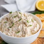 A white bowl filled with creamy smoked salmon dip, garnished with fresh dill and black pepper, sits on a wooden board alongside bread slices, more dill, and a lemon half in the background.