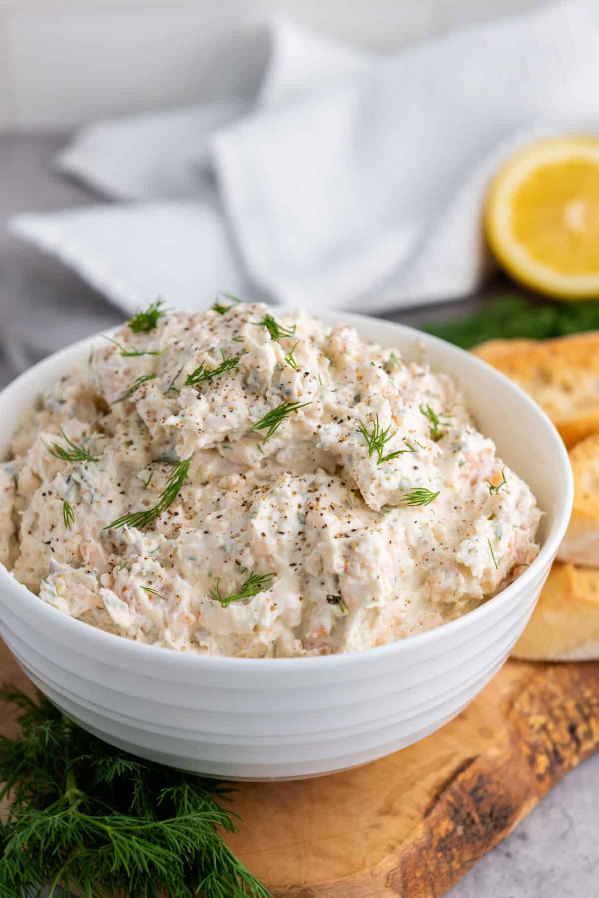 A white bowl filled with creamy smoked salmon dip, garnished with fresh dill and black pepper, sits on a wooden board alongside bread slices, more dill, and a lemon half in the background.