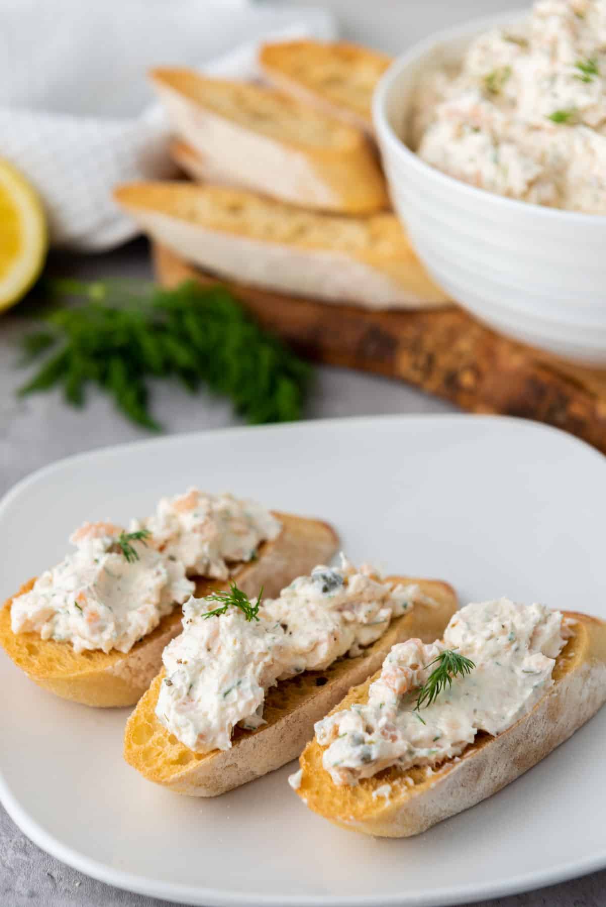 Three pieces of toasted baguette topped with creamy smoked salmon dip and garnished with fresh dill on a white plate, with more bread slices and a bowl of the dip in the background.