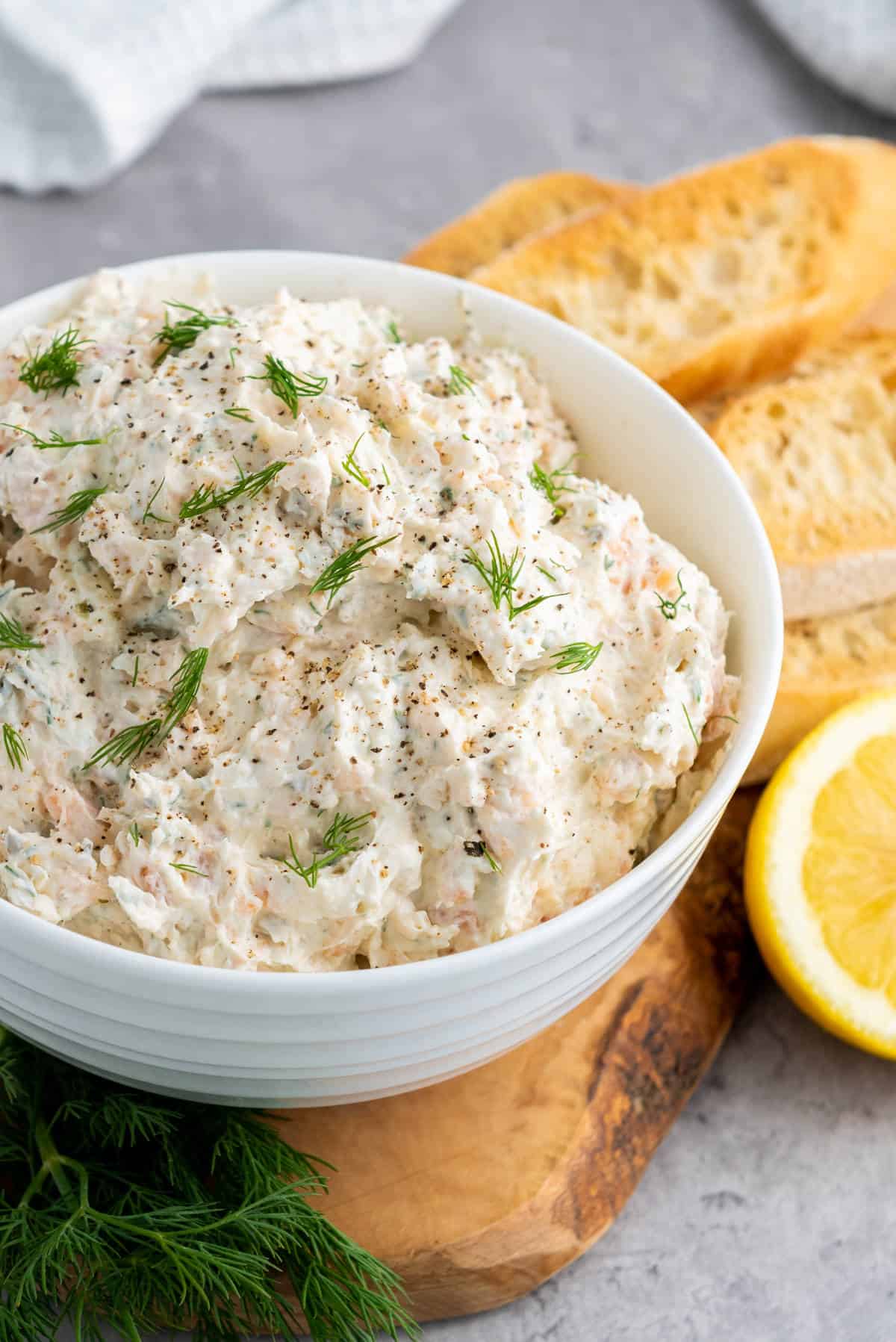 A white bowl filled with creamy smoked salmon dip, garnished with fresh dill and black pepper, sits on a wooden board next to toasted bread slices and a lemon half.
