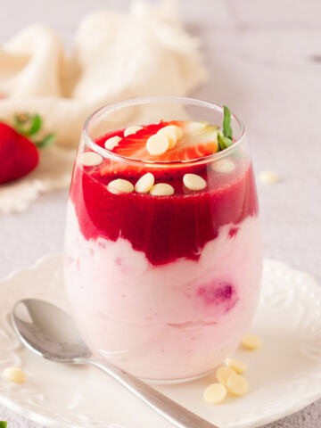 A glass of strawberry mascarpone mousse parfait topped with strawberry sauce, white chocolate chips, and a fresh strawberry slice, served on a white plate with a spoon.