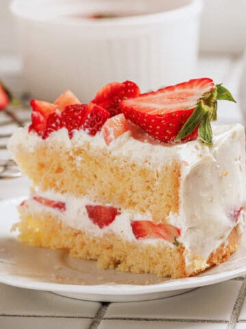 A slice of vanilla sponge cake with whipped cream and fresh strawberries between the layers and on top, served on a white plate with a bite taken out.