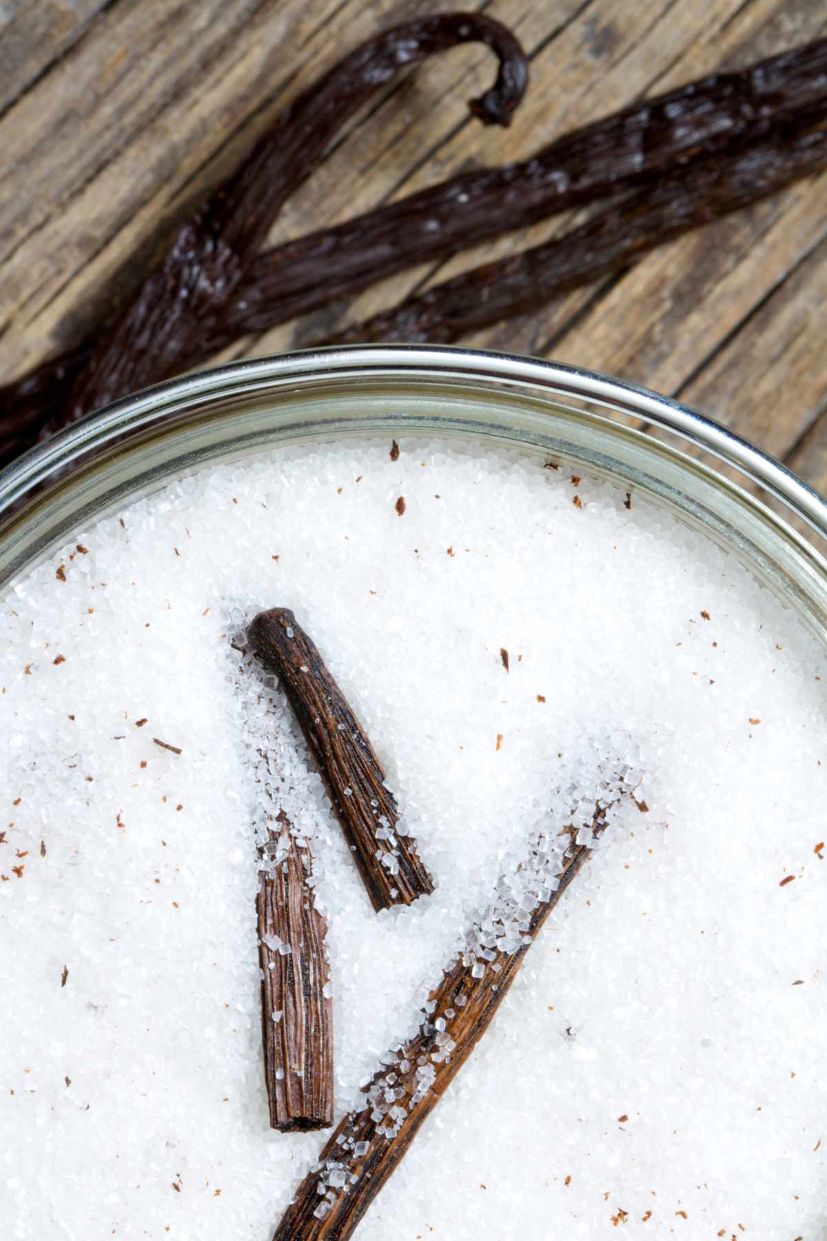 Close-up of a glass jar of vanilla sugar with two vanilla bean pieces on top, and whole beans on a rustic wooden background.