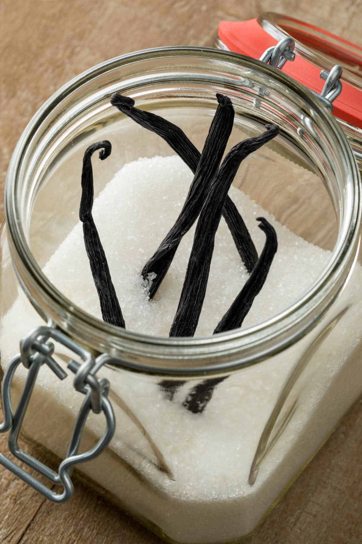 Glass jar of vanilla sugar with whole vanilla beans in white sugar on a wooden surface.