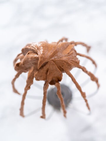 A whisk attachment covered in whipped chocolate ganache, held above a blurred white marble surface.