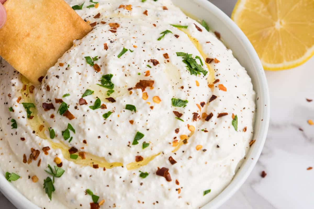 A bowl of creamy whipped feta cheese dip garnished with chopped parsley, crushed red pepper, black pepper, and a drizzle of olive oil, with a pita chip being dipped and a lemon slice in the background.