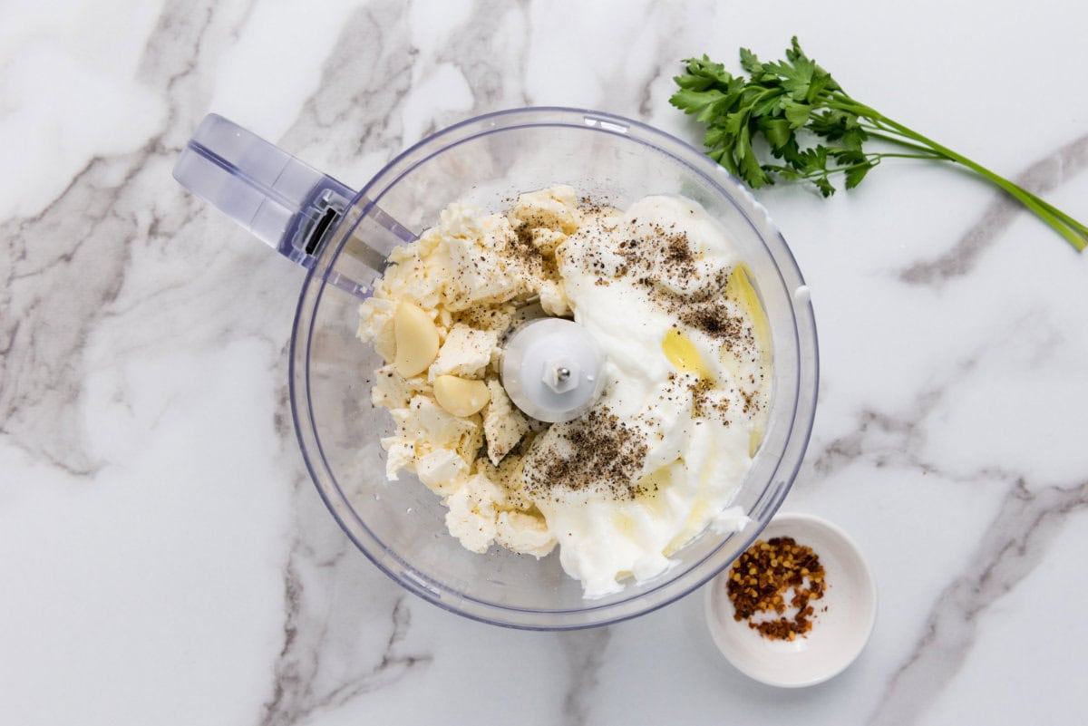 A food processor bowl filled with ricotta cheese, garlic cloves, olive oil, and black pepper sits on a marble surface, ready for blending into a creamy whipped feta cheese dip. Nearby are a sprig of fresh parsley and a small bowl of red pepper flakes.