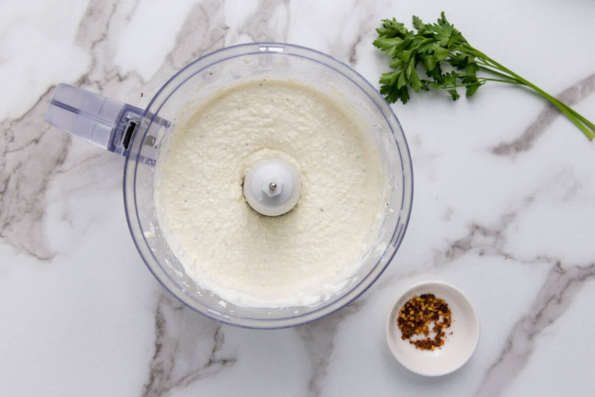 A food processor filled with whipped feta cheese dip sits on a marble countertop. Nearby are a sprig of fresh parsley and a small white bowl containing red pepper flakes, ready to garnish the creamy mixture.
