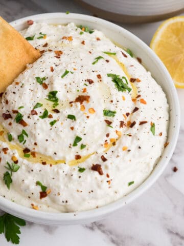 A bowl of whipped feta dip topped with chopped herbs, red pepper flakes, black pepper, and a drizzle of olive oil. A pita chip is being dipped in. A lemon slice and fresh parsley are nearby.