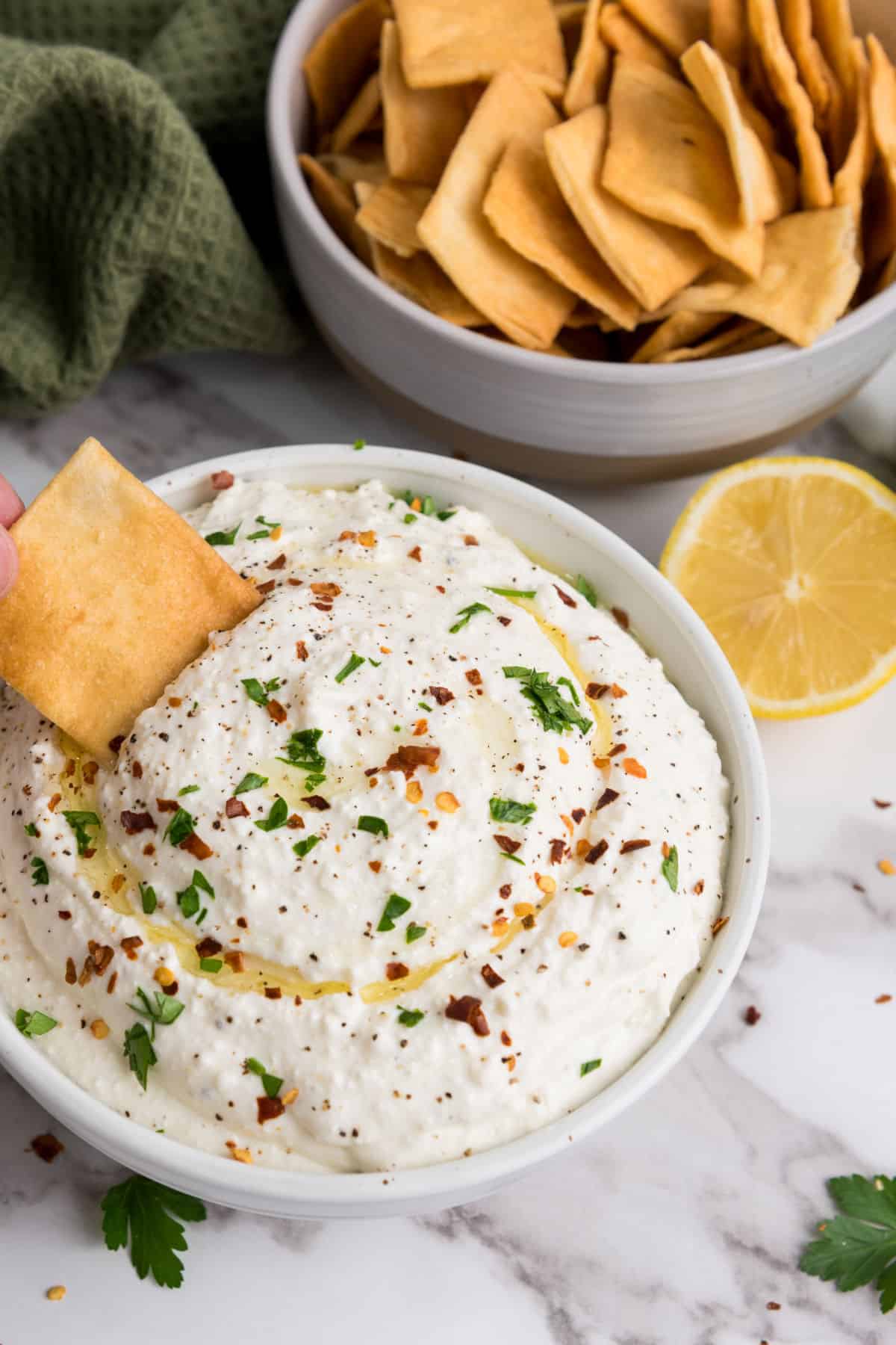 A bowl of whipped feta cheese dip garnished with chopped parsley, pepper, and red pepper flakes, with a pita chip being dipped. In the background, there’s a bowl of pita chips, a lemon half, and a green cloth.