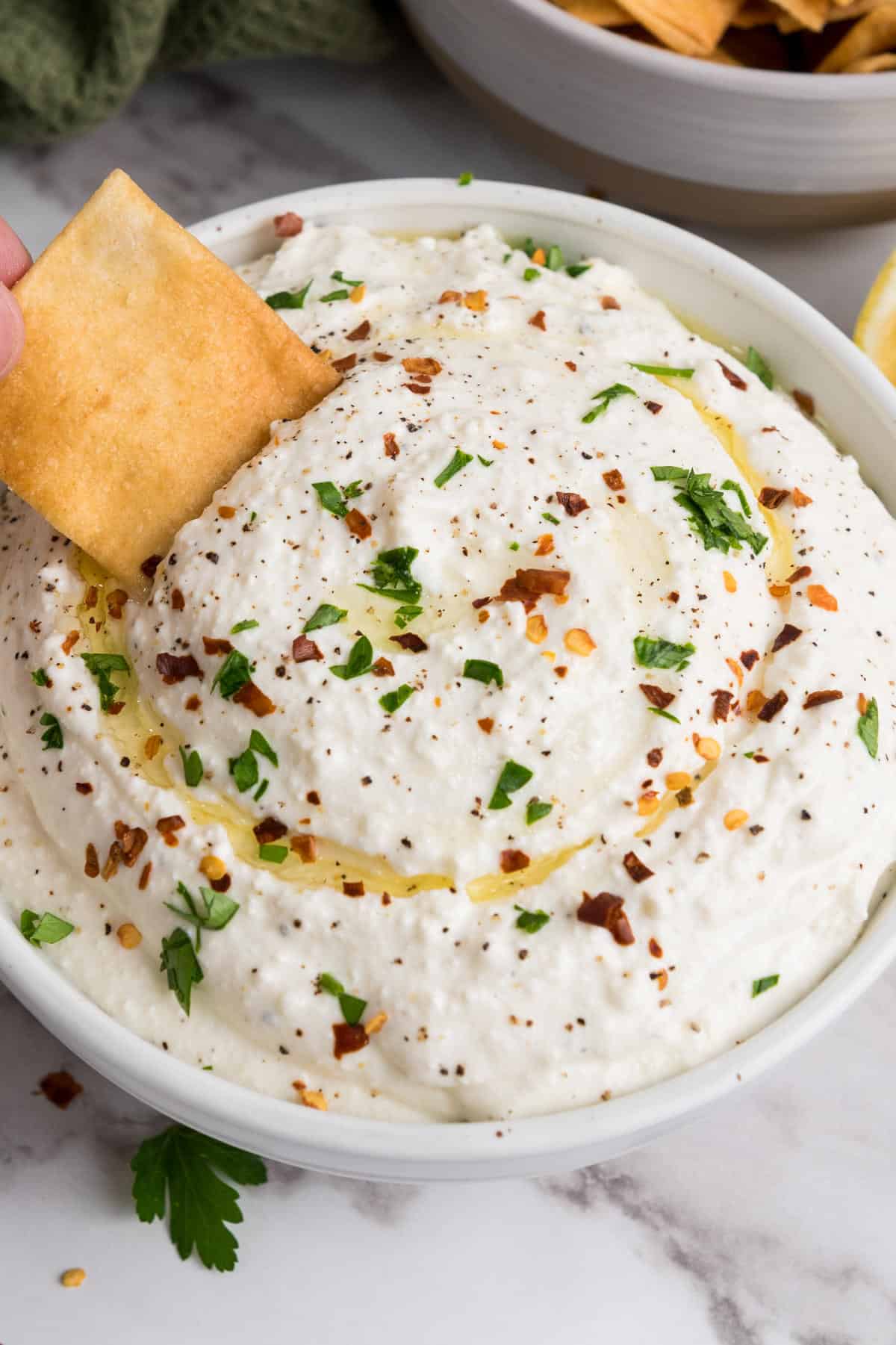 A bowl of whipped feta cheese dip garnished with chopped herbs, red pepper flakes, black pepper, and a drizzle of olive oil. A hand dips a square pita chip into the dip. Chips and a lemon are in the background.