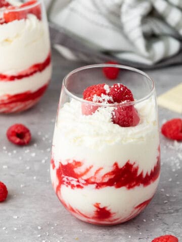 A glass filled with layers of creamy white chocolate raspberry mousse and vibrant raspberry sauce, topped with fresh raspberries and white chocolate shavings. Another similar glass and scattered raspberries are in the background.