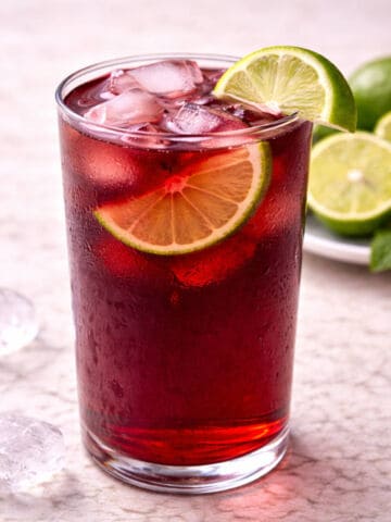 A tall glass of iced red drink garnished with lime slices, set on a light surface with ice cubes, whole and sliced limes, and mint leaves in the background.