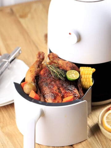 A roasted whole chicken garnished with lime and herbs sits in an air fryer basket, surrounded by pieces of corn and vegetables on a wooden table with plates, tongs, and sliced lemon nearby.