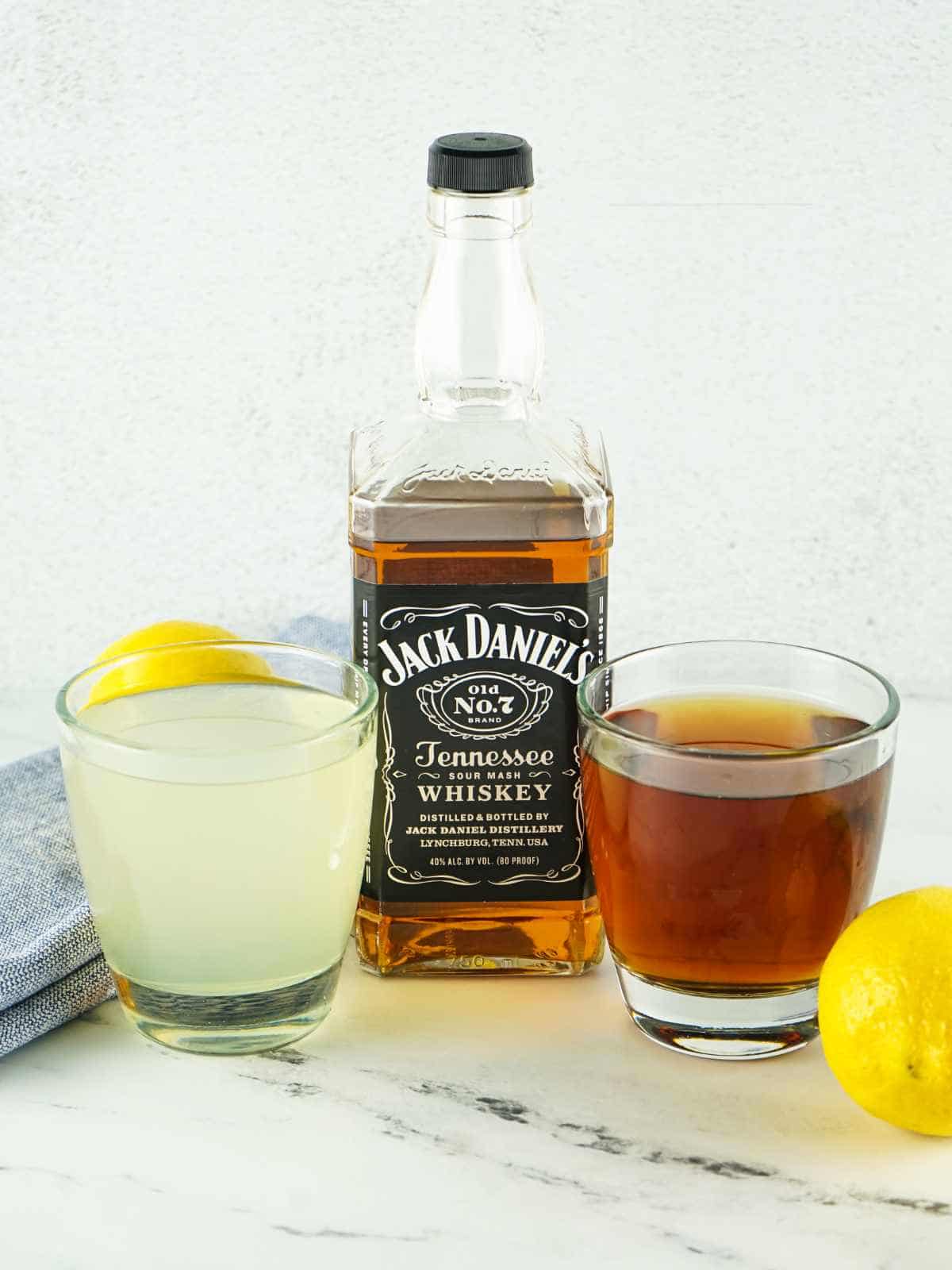 A bottle of Jack Daniel’s Tennessee Whiskey is placed between two glasses, one filled with a yellow lemonade and the other with a dark iced tea. Lemons and a blue cloth are nearby on a light background.