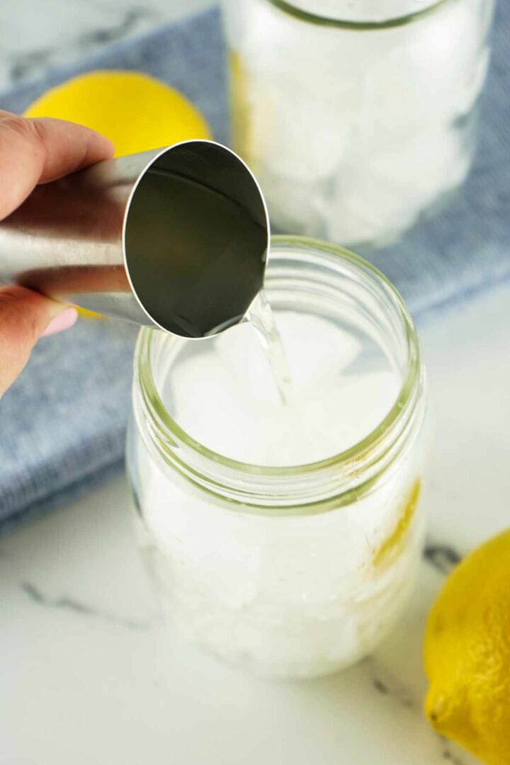 A hand pours a clear liquid from a metal jigger into a mason jar filled with ice, with a lemon and another glass in the background on a blue cloth.