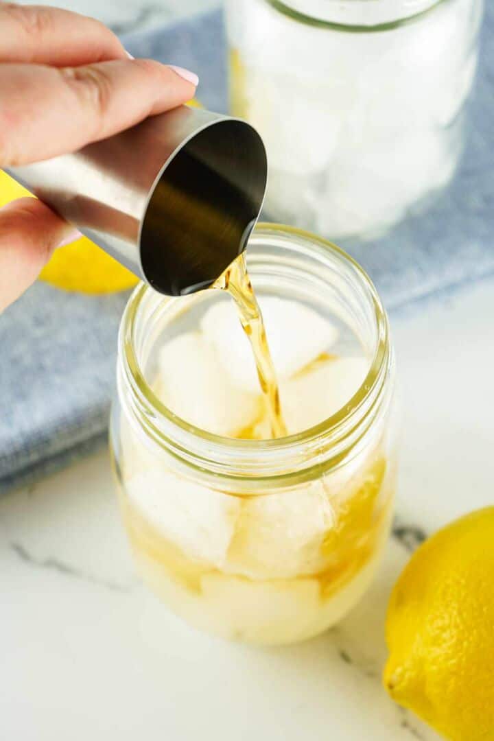 A hand pours a golden liquid from a metal jigger into a mason jar filled with ice, with a lemon and a blue cloth in the background.
