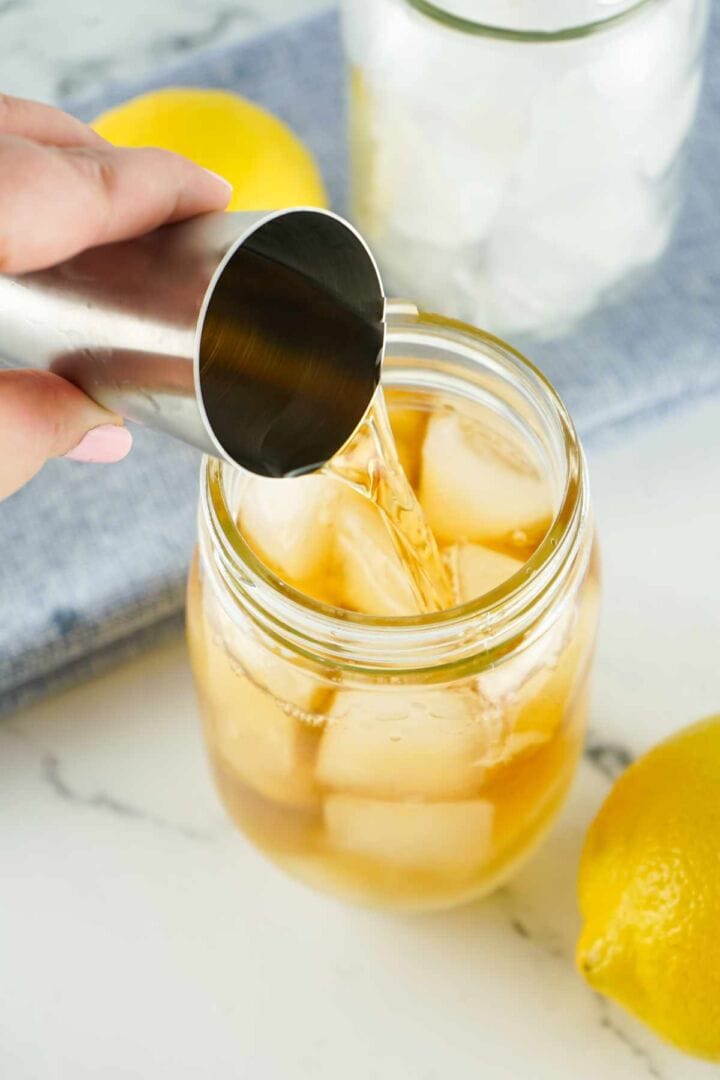 A hand pours a light brown liquid from a metal jigger into a mason jar filled with ice cubes. Lemons and a glass of ice are nearby on a white countertop.