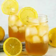 Two mason jars filled with iced tea and ice cubes, each garnished with a lemon slice on the rim. Fresh lemons and a checkered cloth are in the background.