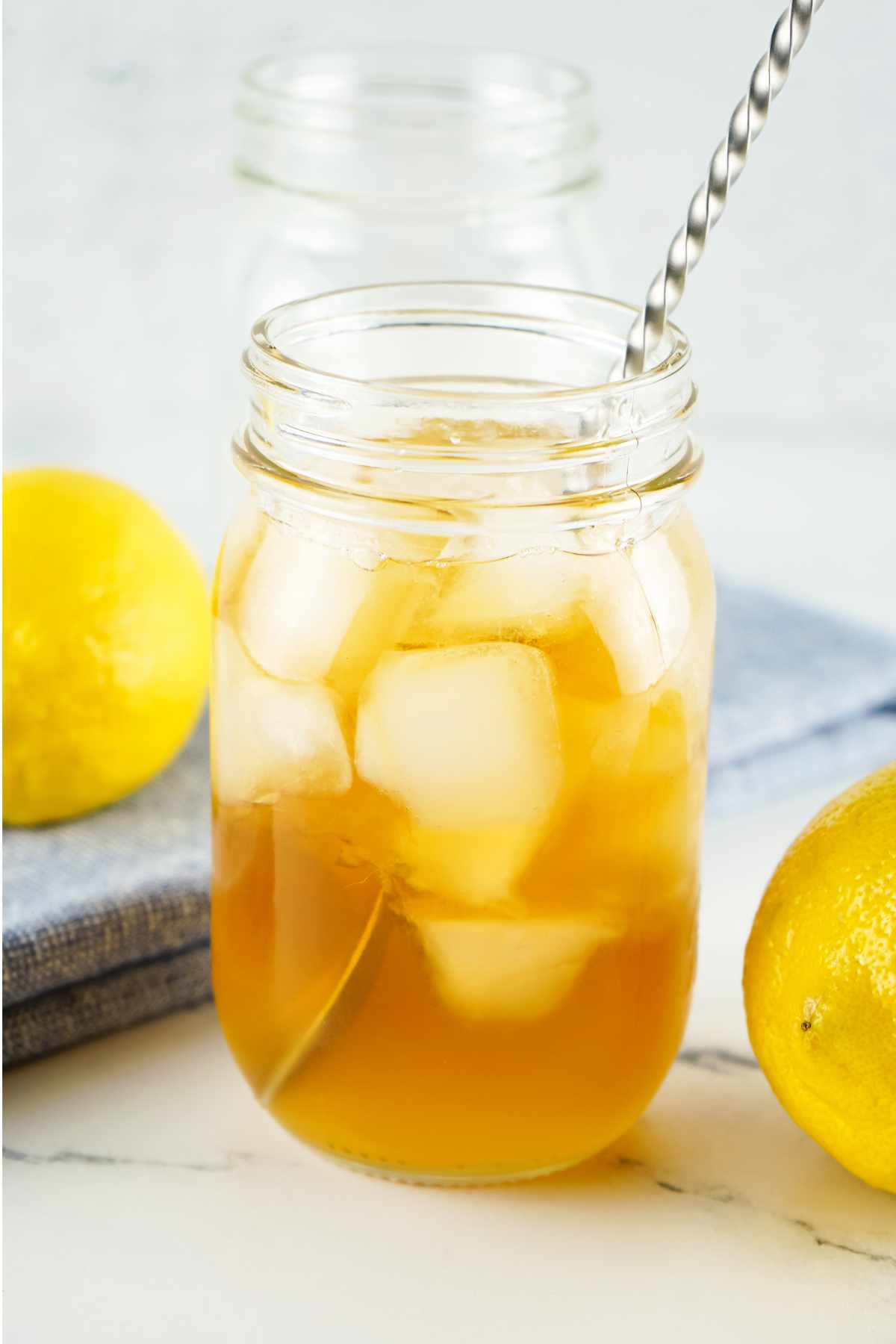 A glass mason jar filled with iced tea and ice cubes, with a metal straw, sits on a white surface next to two whole lemons and a folded blue cloth.