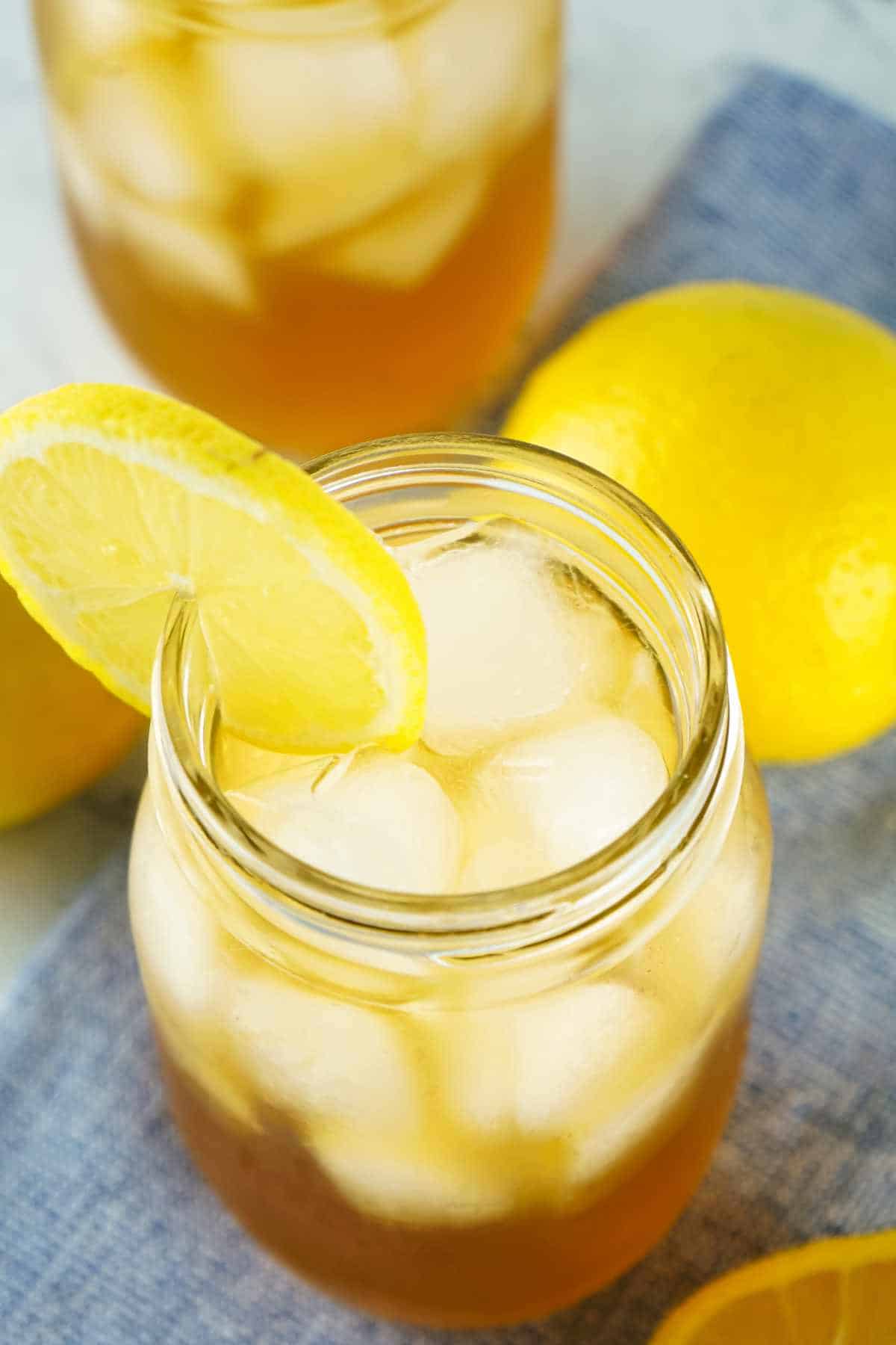 A mason jar filled with iced tea and ice cubes, garnished with a lemon slice, sits on a blue cloth with whole lemons nearby. Another jar of iced tea is partially visible in the background.