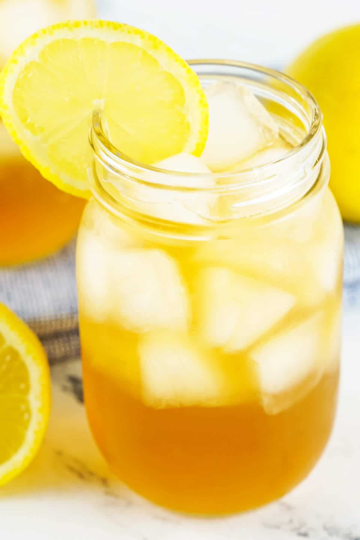 A mason jar filled with iced tea and ice cubes, garnished with a lemon slice on the rim. A whole lemon and another lemon slice are visible in the background.