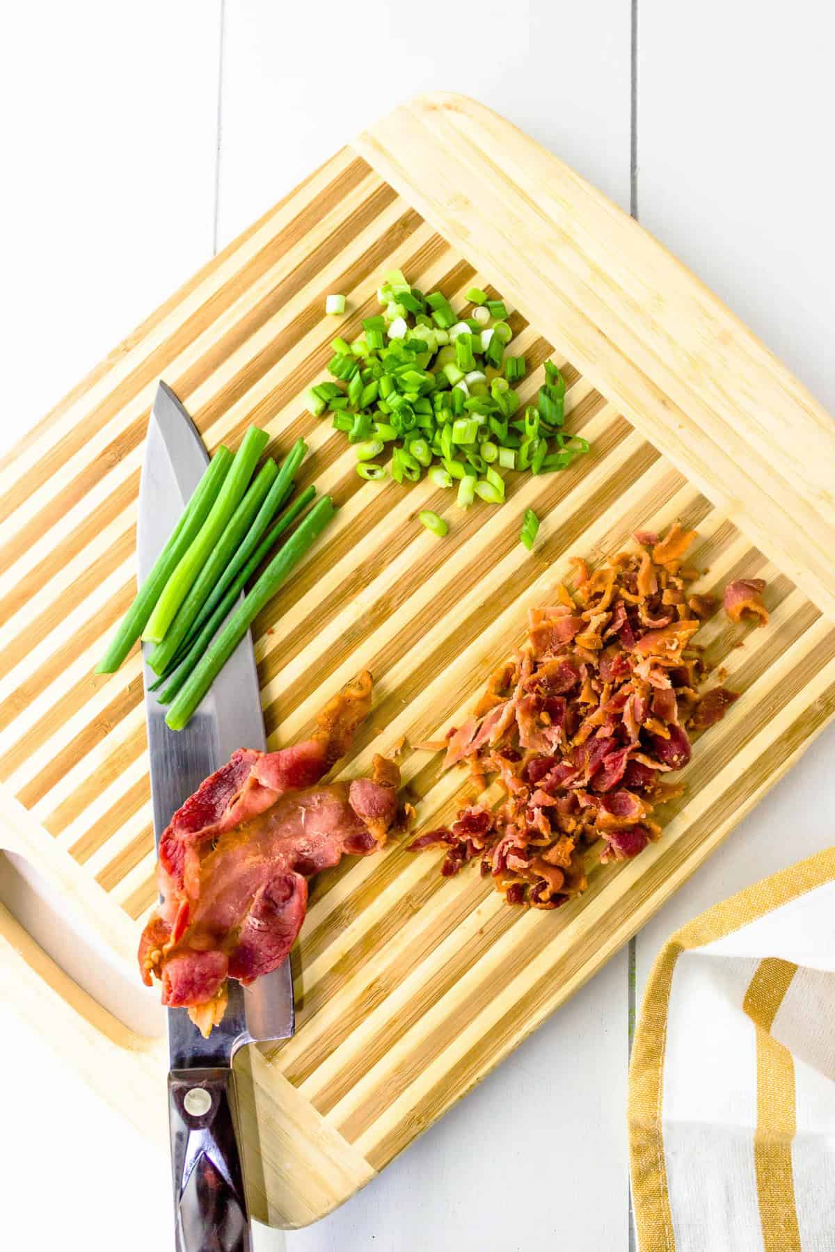 A wooden cutting board with chopped green onions, whole green onions, cooked bacon strips, crumbled bacon, and a chef’s knife, next to a white and yellow kitchen towel.