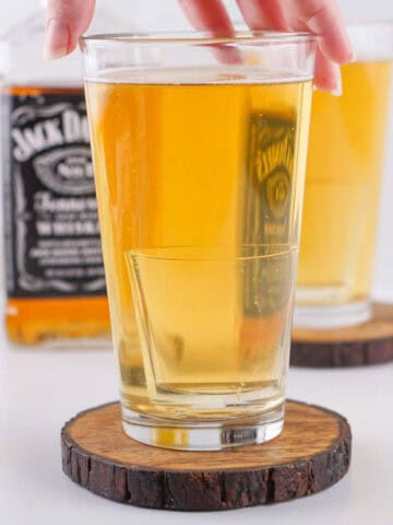 A hand holds a glass of light golden drink on a wooden coaster, with another similar glass and a bottle of Jack Daniel's whiskey in the background.