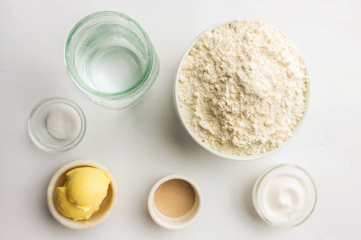 A top-down view of baking ingredients on a white surface: a bowl of flour, a glass of water, small bowls of salt, softened butter, yeast, and sugar.