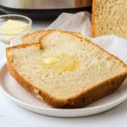 A thick slice of homemade bread with melted butter on top sits on a white plate. In the background are a bowl of butter and more bread on parchment paper.