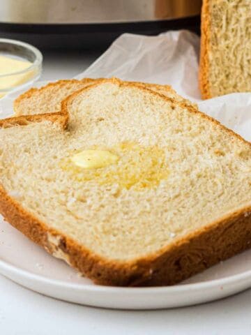 A thick slice of homemade bread with melted butter on top sits on a white plate. In the background are a bowl of butter and more bread on parchment paper.