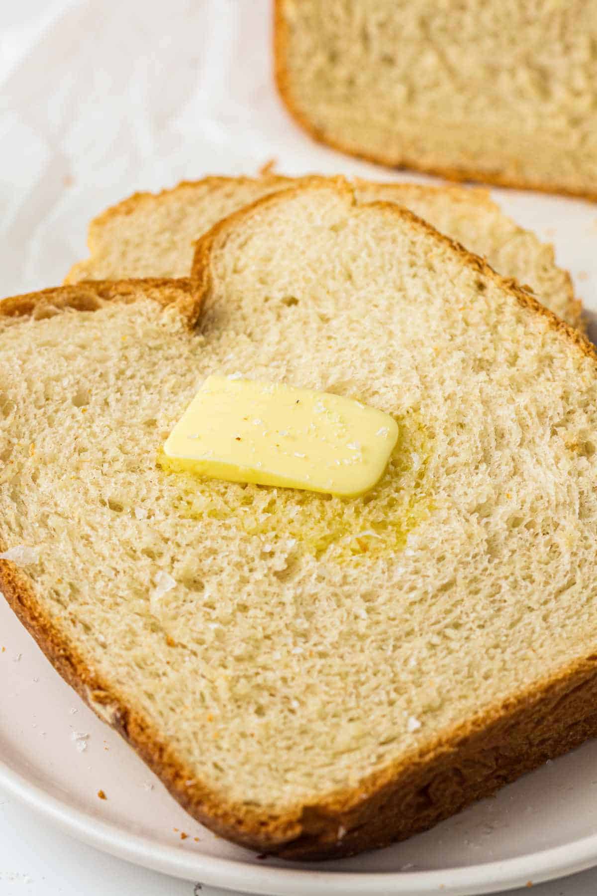 A slice of white bread with a pat of butter melting on top, placed on a white plate with another slice of bread partially visible in the background.