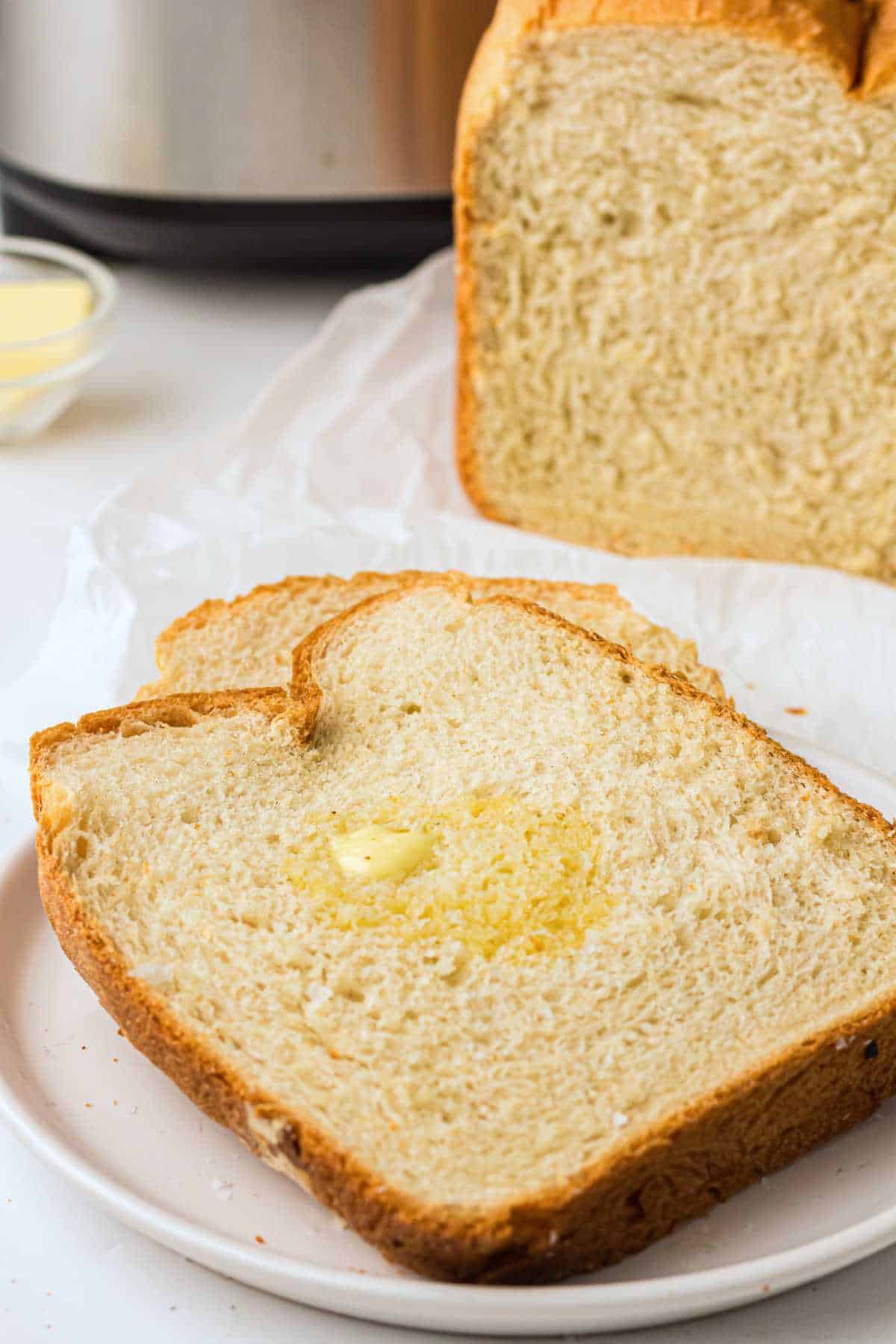 A close-up of two slices of homemade bread on a white plate, with a pat of melted butter on one slice. In the background, a larger loaf of bread sits on parchment paper.