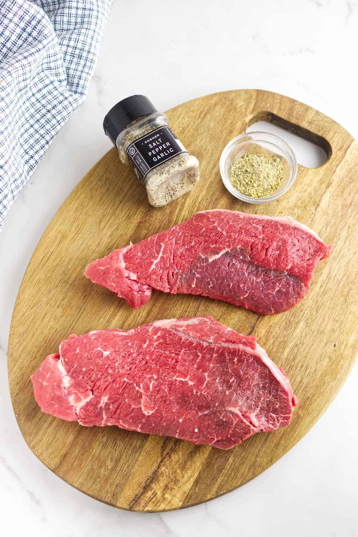 Two raw beef steaks on a wooden cutting board with a small container of dried seasoning and a jar labeled “garlic.” A blue and white towel is partially visible in the upper left corner.