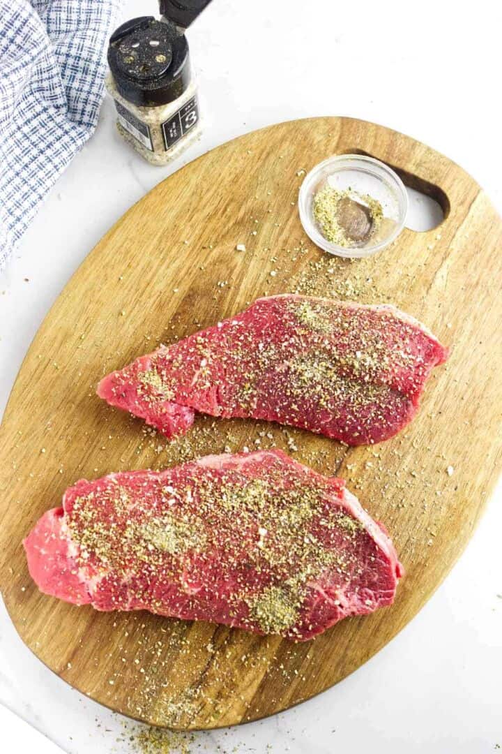 Two raw steaks seasoned with spices are placed on a wooden cutting board. A small container of seasoning and a pepper grinder are nearby, with a blue and white cloth in the corner.