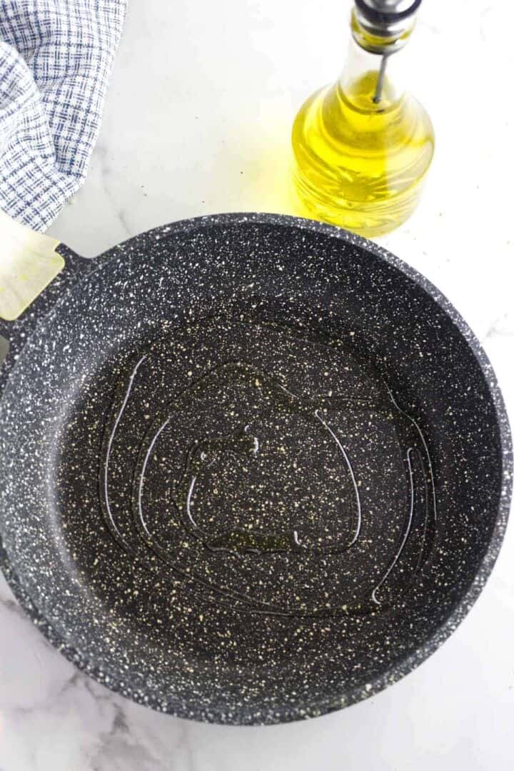 A black speckled frying pan with a swirl of olive oil inside sits on a marble countertop. Nearby, there is a glass bottle of olive oil and a blue-and-white checkered cloth.